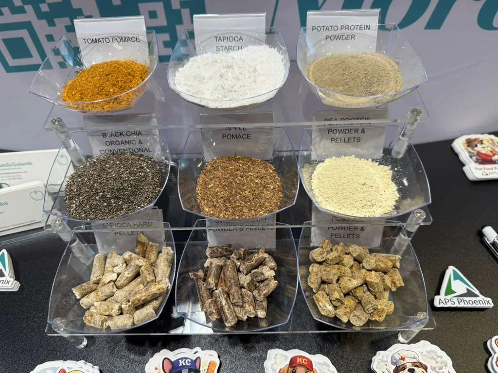 Ingredient sample cups on the APS Phoenix Booth 1026 table at Petfood Forum 2026: tomato pomace, tapioca starch, potato protein, black chia organic, apple pomace, pea protein, tapioca pellets, sweet potato, pumpkin powder.